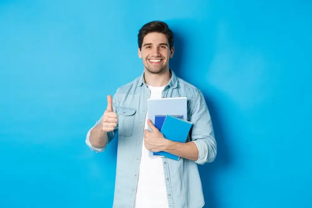 young-man-student-with-notebooks-showing-thumb-up-approval-smiling-satisfied-blue-studio-background